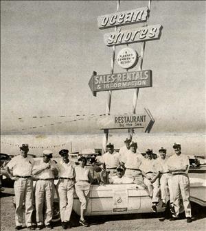 Eleven men and women in white shirts and matching caps sit and stand around a vintage car underneath a large sign that says Ocean Shores Sales Rental Information Restaurant by the Sea. Image is black and white. 