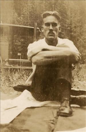 A man sits on the ground with his arms wrapped around one knee in front of an early 20th century car and a forest. 