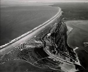 An aerial view of a long peninsula that stretches into the ocean, a bay behind the peninsula, and roads crisscrossing the peninsula. Image is black and white. 