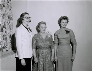 Three women in dresses stand in a corner smiling past the camera. 