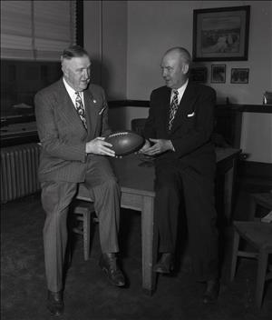 Two white man in suits lean against a table while one hands a football to the other 