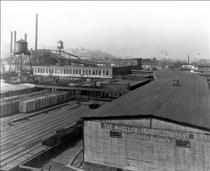 Wooden factory buildings and smokestacks with railroad tracks running between them. 
