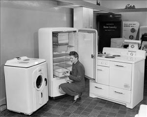 A smiling woman in a dress crouches in front of an empty refrigerator with its door open. Other white appliances are in the room. 