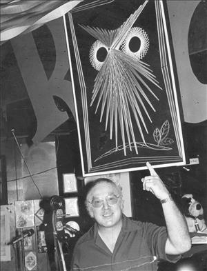 A white man points to a framed artwork of an owl face above him. 