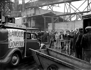 Men in work clothes stand around a man in a suit speaking at a microphone. They are outside a factory. A car parked beside them has a sign that says Langlie for Governor. 