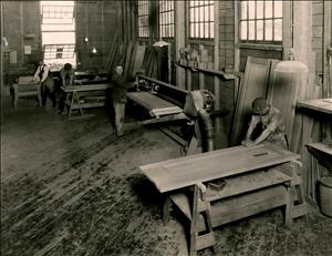 Men lean over workbenches in a wooden warehouse with large windows. 