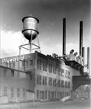 A three story warehouse building beneath a wooden water tower with smokestacks in the background. The word Plywood is painted on the side of the warehouse. 