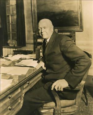 A white man in a suit sits at a large desk covered in papers.