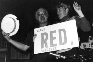 A white man holds up a straw hat next to a white woman holding a sign that says Elect Red WhoCrat. There is a saxophone in the foreground. 