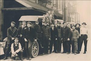 A large group of young people stand around an early twentieth century automobile. 