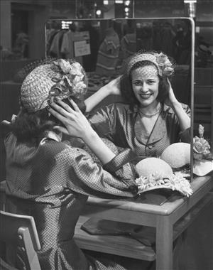 A smiling woman is reflected in a mirror with her hands holding a straw hat to her head. 