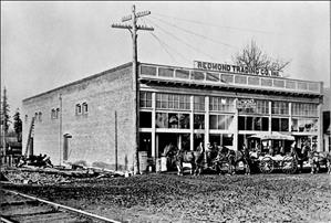 A one story storefront with horses and a horse drawn carriage in front. Train tracks pass by on one side of the building. A sign on the roof says Redmond Trading Post and a sign in the window says Redmond Post Office. 
