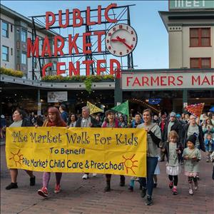 Adults and children march behind a banner that says Pike Market Walk for Kids. They are walking beneath a large neon sign that says Public Market Center with a clock next to it