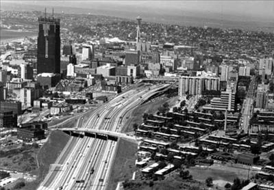 a multi lane freeway curves through a city. on one side there are skyscrapers. There are two overpasses over the freeway. 