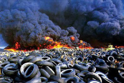 Dramatic photo of junked tires stacked as far as the eye can see at a dump in Everett, with bright orange and yellow flames and oily black smoke billowing in the near distance and blocking the sky. The fire would burn and pollute the air for six months before being extinguished 