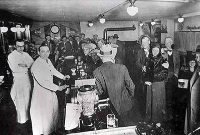 White men in hats sit and stand along a long bar. Two bartenders in white coats stand behind the bar