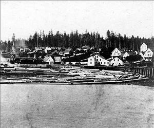 Small wooden buildings along the shore of a bay. Rafts of logs float in the water. Dense forest in the background. 