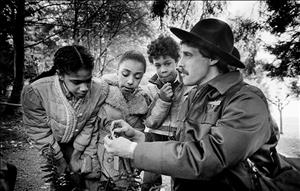 A man in a ranger hat and jacket holds something small in his hands. Three youth of color lean in to see what is in the rangers hands. They are in the forest. 