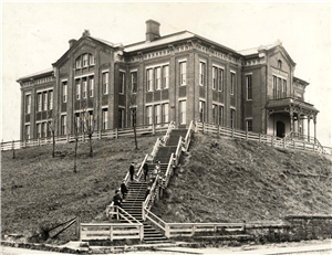 A large brick school building at the top of a long flight of stairs. Children in early twentieth century clothing stand on the steps. 