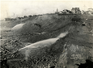 Men direct large streams of water towards a bluff upon which there are wooden buildings and houses. 