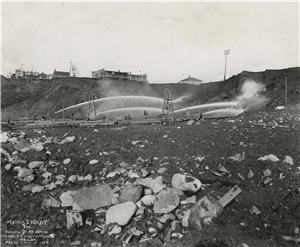 Men direct large streams of water towards a bluff upon which there are wooden buildings and houses. 