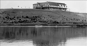 Black and white image of a lake with a large building on the hill behind it. 