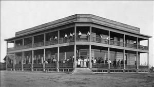 Black and white image of a large two story building with men and women on the wrap around porch and balcony