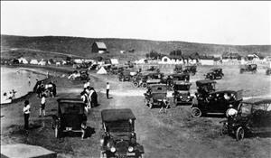 Black and white image of cars parked along the lakeshore. People on the beach and houses in the background.