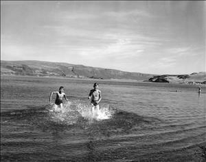 Black and white photo of two boys in swim trunks running out of a lake. 