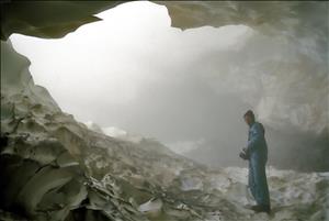 A person in a snowsuit stands inside a cave of ice. The entrance to the cave is filled with steam. 