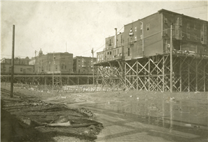 Old wooden buildings on tall wooden pilings or scaffolding. Beneath them is mud. 