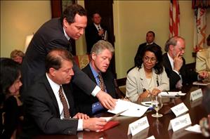 Men and women in suits sit around a wooden table. A man leans between two people to place a paper on the table. 