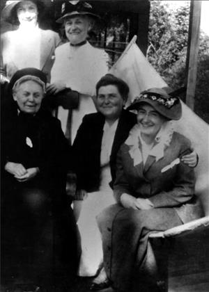 Five women in dresses and hats sit and stand close to each other for the photograph. 